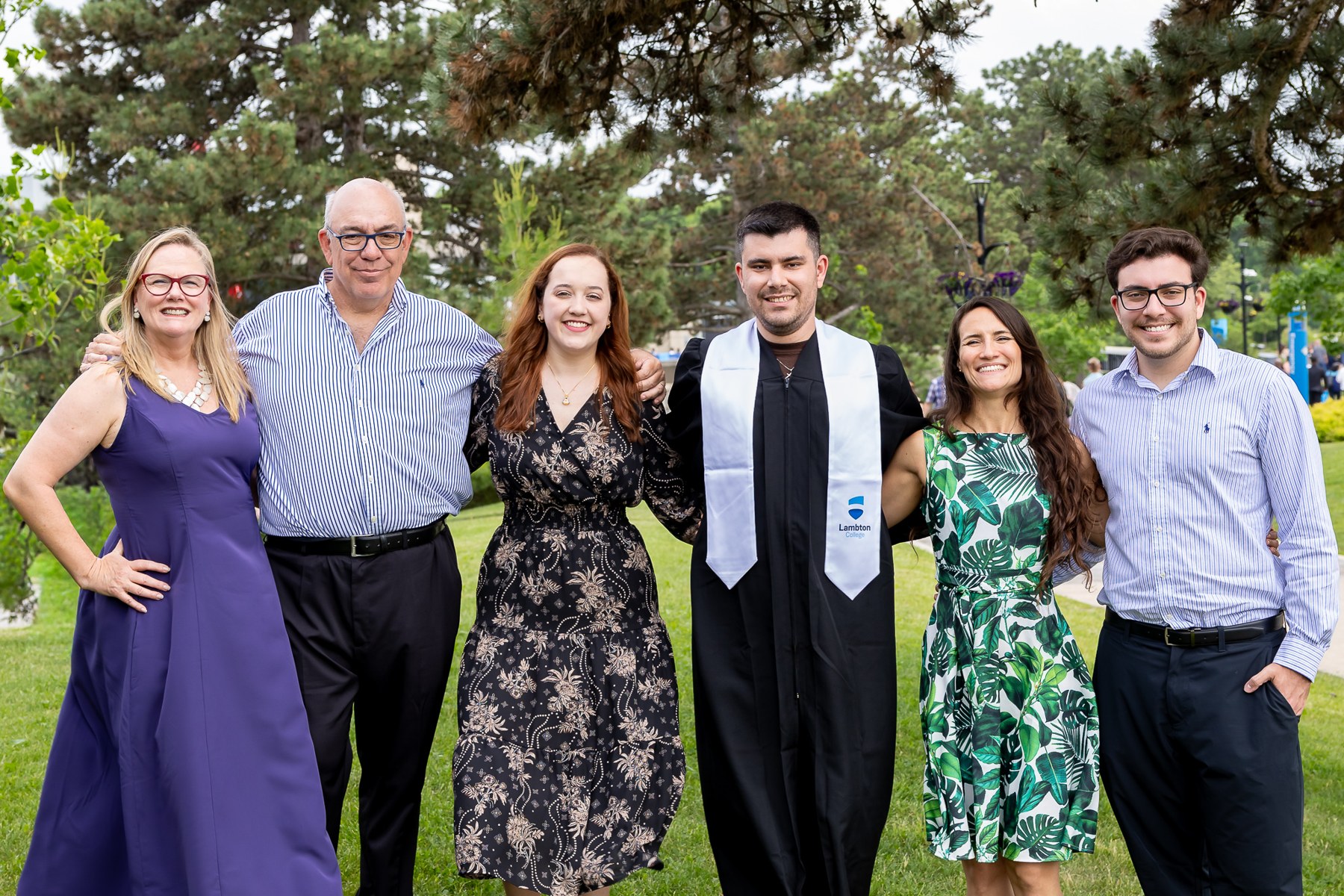 Graduate standing with family outdoors beneath leafy trees