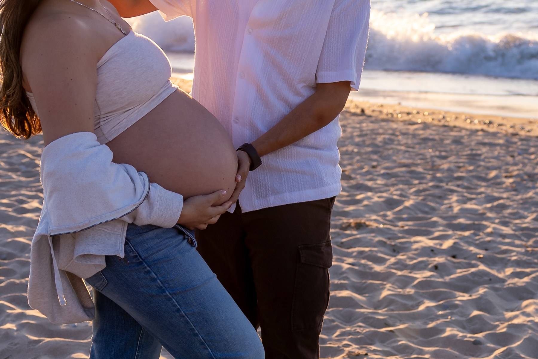 Close-up of a couple’s hands resting together on a baby bump at the beach