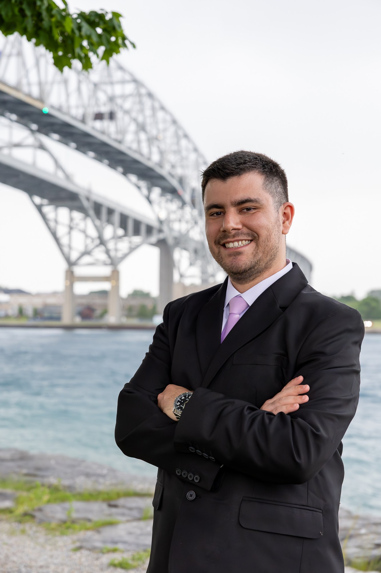 Graduate in a suit standing with arms folded beside a bridge