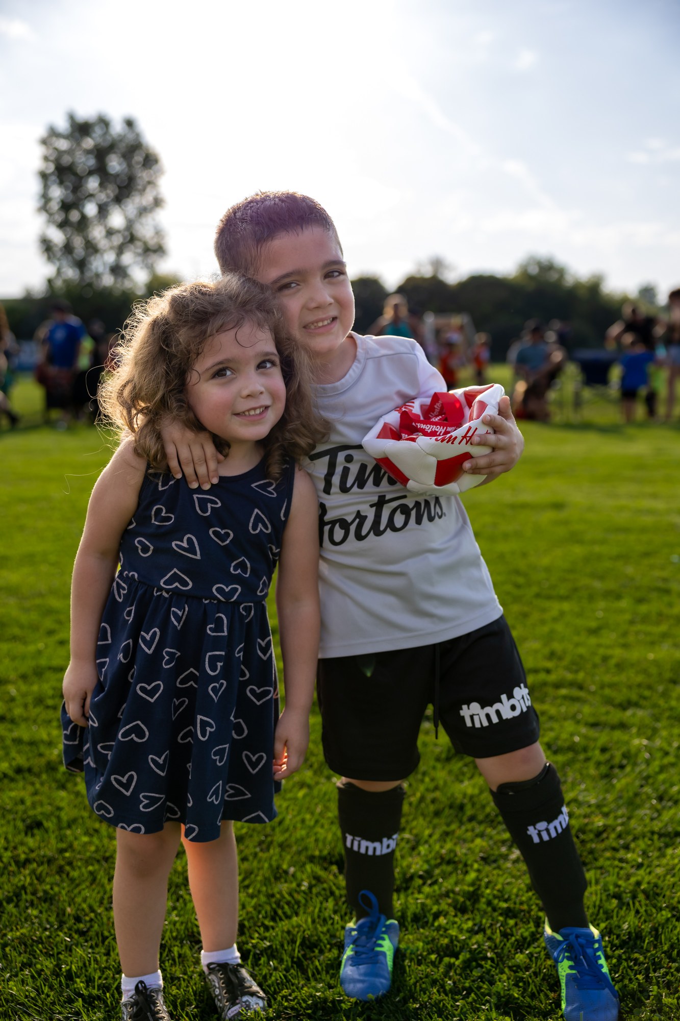 Two children standing together on the grass after a soccer game, one holding a ball